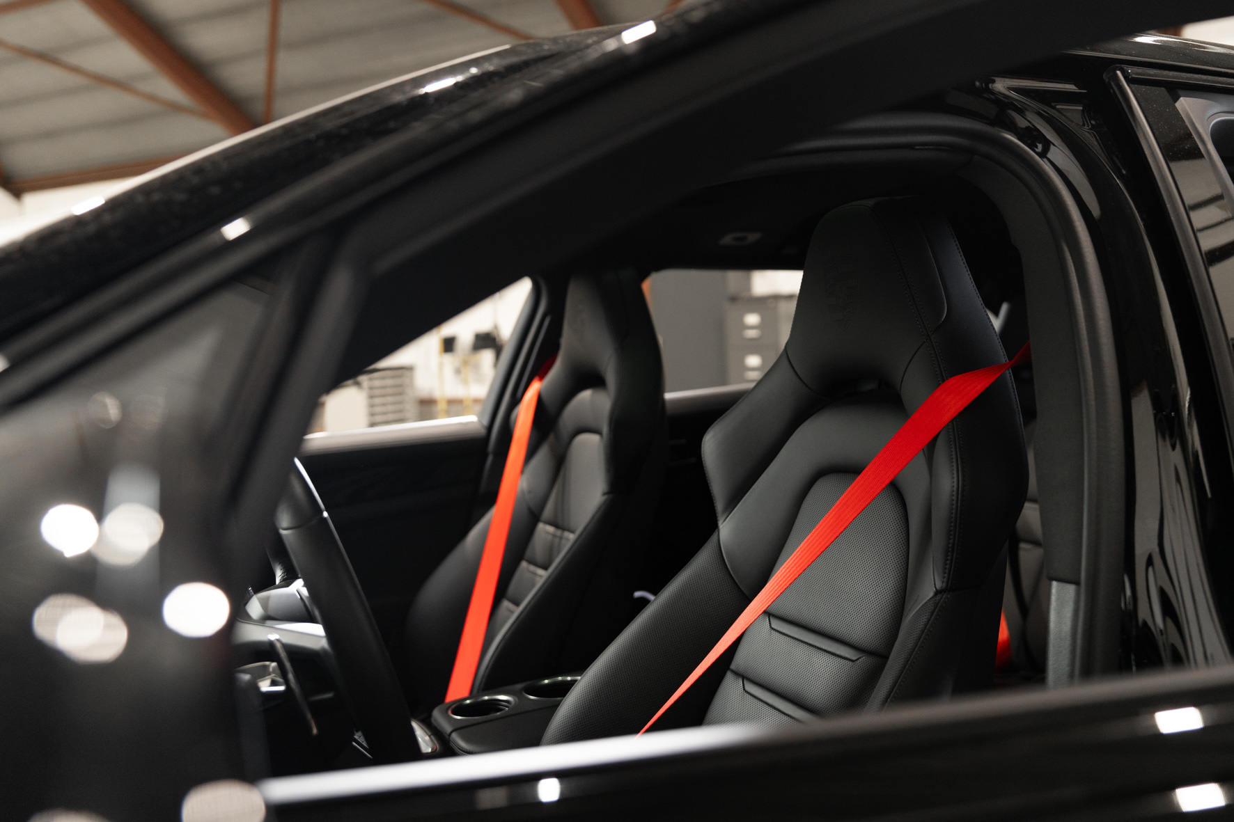 Zijaanzicht van twee sportstoelen in een zwarte auto met rode autogordels, gefotografeerd in een werkplaats.