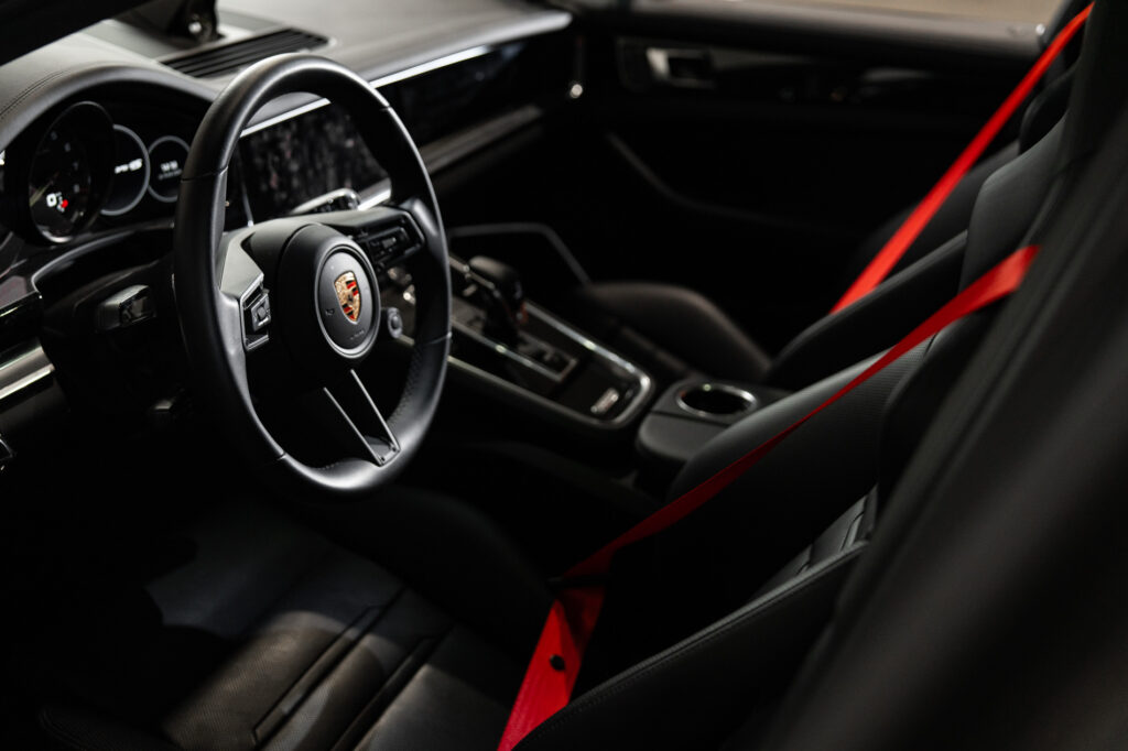 Interieur van een Porsche met rood-zwarte sportstoelen en rode autogordels, gefocust op stuur en middenconsole.