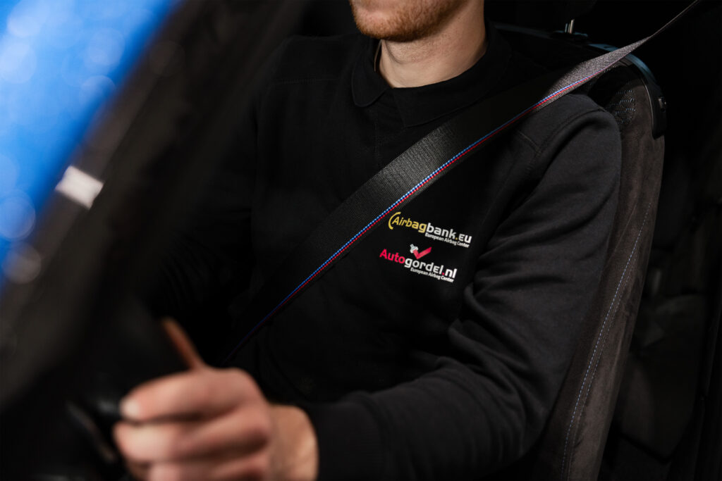Medewerker van Autogordel.nl draagt een zwarte trui met bedrijfslogo’s en een BMW M-stijl autogordel tijdens het autorijden.